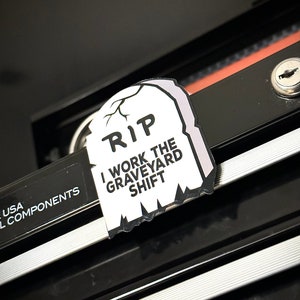 May include: A white and gray tombstone-shaped sticker with the text "RIP I WORK THE GRAVEYARD SHIFT." The sticker is placed on a black surface. The sticker has a jagged edge design.