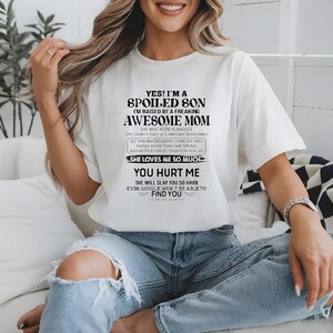 K&ouml;nnte beinhalten: Wei&szlig;es T-Shirt mit schwarzem Text: "YES! I'M A SPOILED SON I'M RAISED BY A FREAKING AWESOME MOM." Das Shirt hat auch zus&auml;tzlichen Text darunter. Das Model tr&auml;gt zerrissene blaue Jeans.