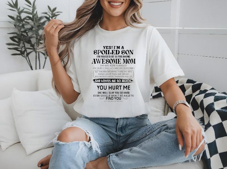 K&ouml;nnte beinhalten: Wei&szlig;es T-Shirt mit schwarzem Text: "YES! I'M A SPOILED SON I'M RAISED BY A FREAKING AWESOME MOM." Das Shirt hat auch zus&auml;tzlichen Text darunter. Das Model tr&auml;gt zerrissene blaue Jeans.