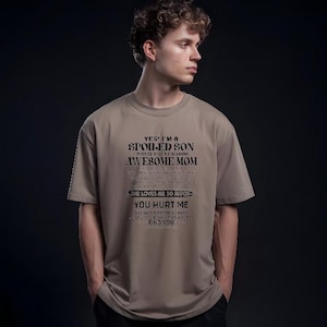 K&ouml;nnte beinhalten: Ein beiges T-Shirt mit dem Text "YES, I'M A SPOILED SON RAISED BY A MEANING AWESOME MOM. SHE LOVES ME SO MUCH. YOU HURT ME, SHE'LL FIND YOU." Das T-Shirt wird von einer Person getragen.