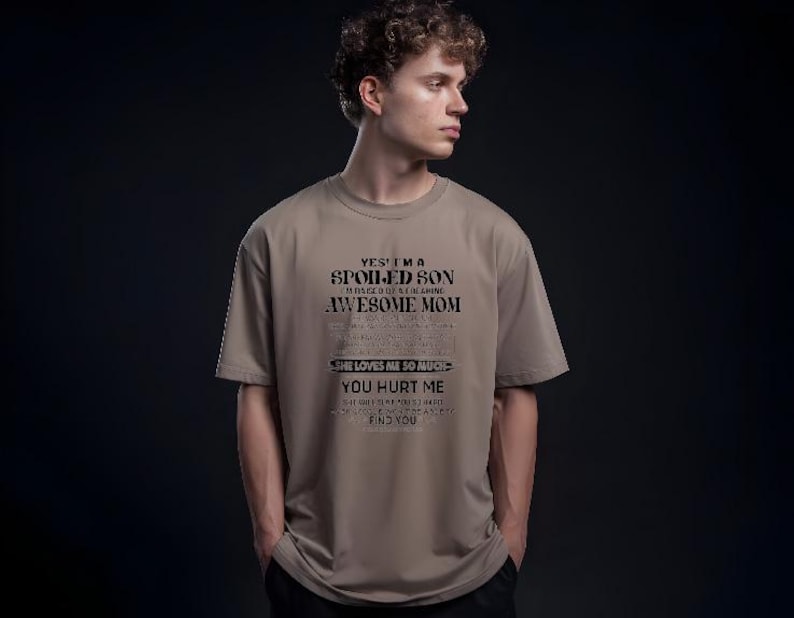 K&ouml;nnte beinhalten: Ein beiges T-Shirt mit dem Text "YES, I'M A SPOILED SON RAISED BY A MEANING AWESOME MOM. SHE LOVES ME SO MUCH. YOU HURT ME, SHE'LL FIND YOU." Das T-Shirt wird von einer Person getragen.