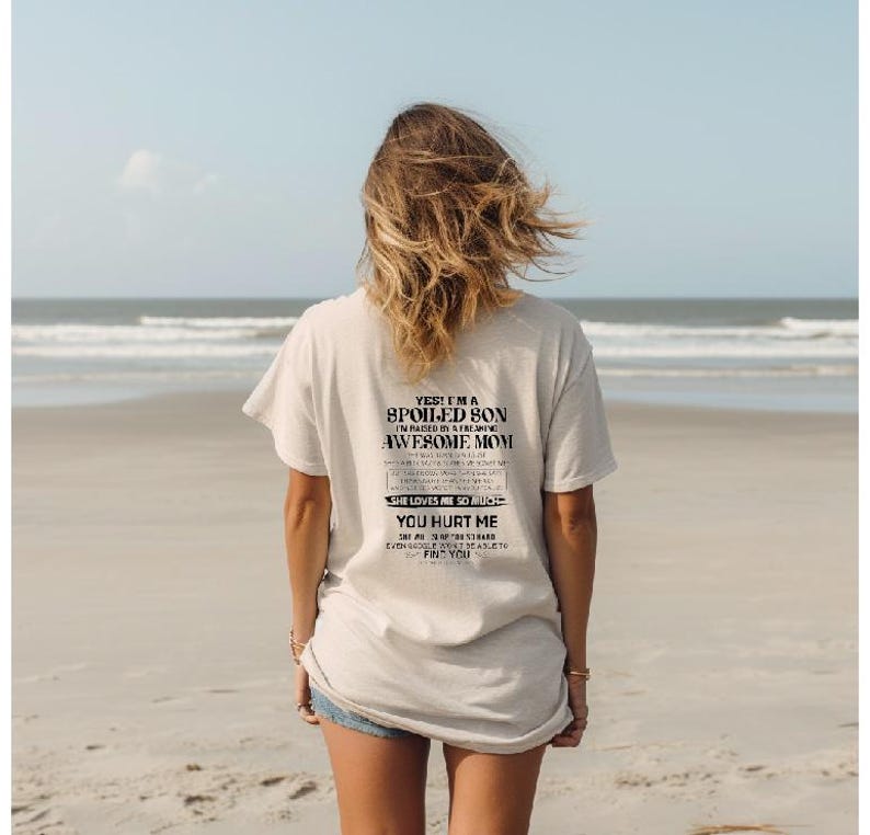 K&ouml;nnte beinhalten: Hellbeige T-Shirt mit schwarzem Text auf dem R&uuml;cken. Der Text lautet: "YES! I'M A SPOILED SON IN RAISED BY A FREAKING AWESOME MOM SHE LOVES ME SO MUCH YOU HURT ME EVEN GOOGLE CAN'T EASILY FIND YOU". Die Person steht am Strand.