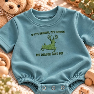 May include: A teal short-sleeved baby bodysuit featuring a green embroidered graphic of a deer with antlers. The text above the deer reads IF IT'S BROWN, IT'S DOWN! and the text below reads MY DIAPER SAYS SO! The garment has a ribbed crew neckline.