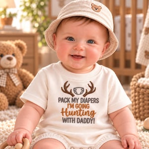 Puede incluir: Un body de bebé de manga corta color crema con un diseño bordado de astas de ciervo marrones, un icono de pañal y el texto "PACK MY DIAPERS I'M GOING Hunting WITH DADDY!" junto a un sombrero de pescador beige a juego con un pequeño parche de oso.