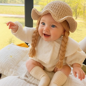 May include: A smiling baby wearing a beige crochet hat with a wavy brim and two long, braided hair extensions. The baby is dressed in a white onesie and socks, sitting on a white blanket with a daisy pattern. The background shows a window with a view of greenery.
