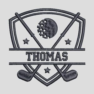 Pode incluir: Design bordado com tema de golfe, apresentando tacos de golfe cruzados, uma bola de golfe e estrelas. O nome "THOMAS" é exibido em letras em negrito no centro. O design é em tons de cinza e preto.