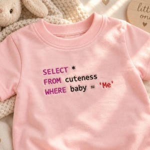 May include: A light pink short-sleeved t-shirt featuring embroidered text that reads SELECT * FROM cuteness WHERE baby = 'Me'. The words SELECT, FROM, and WHERE are in purple, while cuteness, baby, and 'Me' are in black and red.