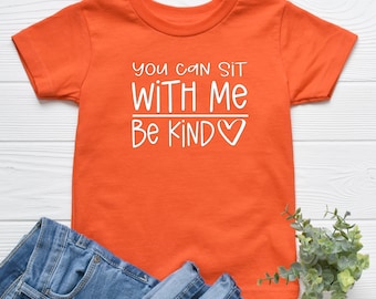May include: An orange t-shirt with the text "You can sit with me, Be Kind" in white. The shirt is paired with blue denim jeans. A small green plant is in the bottom right corner.