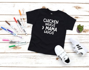 May include: Black t-shirt with white text that reads "Chicken Nugs & Mama Hugs". The shirt is on a white wooden surface with markers and a pair of white sneakers with black stripes.