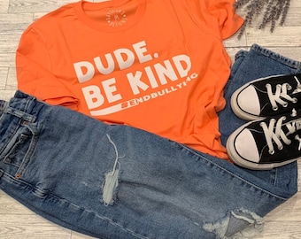 May include: A coral orange t-shirt with the text "DUDE. BE KIND #ENDBULLYING" in white. The shirt is laid on a wooden surface with a pair of blue jeans and black sneakers.