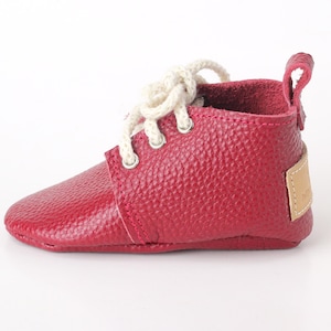 May include: A single red leather baby shoe with white laces. The shoe has a textured surface and a small pull tab at the back. A small tan tag is attached to the side. The shoe is designed for infants.
