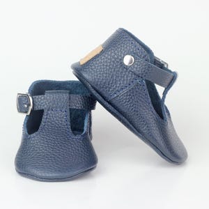 Könnte beinhalten: Ein Paar marineblaue Babyschuhe aus Leder im Mary-Jane-Stil. Die Schuhe haben eine strukturierte Oberfläche, einen Riemen mit Schnalle und einen kleinen Absatz. Sie stehen vor einem weißen Hintergrund.