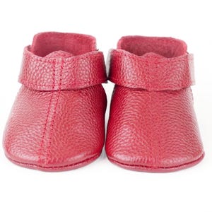 May include: A pair of red baby booties. The booties are made of textured leather and have a folded-over cuff. The stitching is visible in a matching red thread. The booties are designed to be worn on a baby's feet.