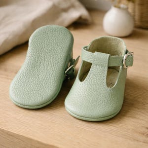 May include: Two light green leather baby shoes with a T-strap design and buckle closure. One shoe is lying flat, showing the sole, while the other is upright. The shoes are on a light wooden surface.