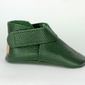 May include: A single, dark green baby shoe made of textured leather. The shoe has a strap with a velcro closure and a soft, green interior. The shoe is designed for infants and toddlers.