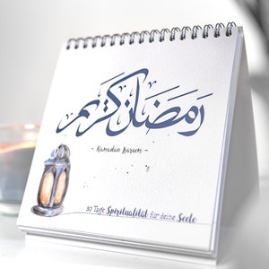 Könnte beinhalten: Ein weißer Tischkalender mit einer blauen arabischen Kalligraphie-Inschrift, die "Ramadan Kareem" sagt, und dem Text "30 Tage Spiritualität für deine Seele". Eine kleine Illustration einer Laterne befindet sich in der linken unteren Ecke.