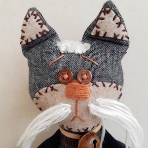 Op de afbeelding: Een grijze en bruine stoffen kattenpop met witte garen snorharen, bruine knoopjesogen en een bruine viltneus. De kat draagt een grijs stoffen jasje met een bruine bel.
