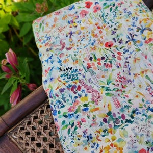 Pode incluir: Um tecido floral com um design em aquarela com uma variedade de flores coloridas em tons de vermelho, amarelo, azul e roxo. O tecido é drapeado sobre uma cadeira de madeira com assento tecido, em um fundo de folhagem verde.