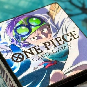 May include: Black card storage box featuring a colorful illustration from the "One Piece Card Game." The box displays the game's logo and character art. The box is designed to hold trading cards.