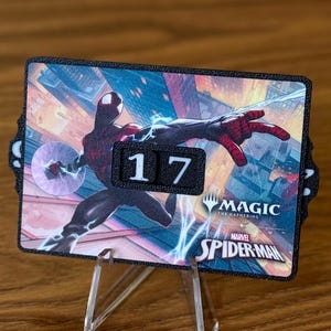Könnte beinhalten: Ein schwarz-roter Spider-Man-Themen-Magic: The Gathering-Kartenhalter mit der Zahl 17. Der Kartenhalter wird von einem transparenten Acrylständer getragen. Die Karte zeigt Spider-Man-Artwork und die Logos Magic: The Gathering und Marvel Spider-Man.