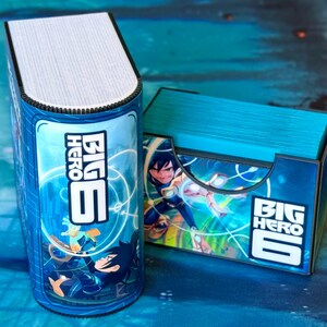 May include: A blue Big Hero 6 themed card holder and a matching card storage box. The card holder is cylindrical and resembles a book, while the box is rectangular. Both feature character illustrations and the Big Hero 6 logo.
