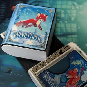 Może przedstawiać: Zestaw gier fantasy z ilustracją rudego lisa na niebieskim tle. Pudełko z grą ma kształt książki z napisem "Winterspell" na okładce. Zawiera kostki i elementy gry.
