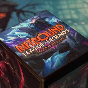 Puede incluir: Una caja negra con el logotipo "Riftbound League of Legends Trading Card Game" en naranja y blanco. La caja tiene un diseño colorido con arte de fantasía azul y morado. La caja es probablemente para guardar cartas coleccionables.