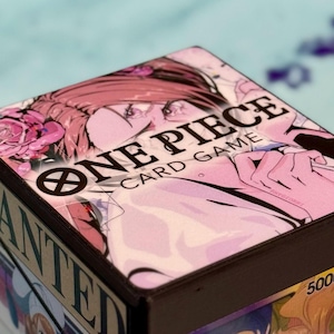 Op de afbeelding: Een One Piece kaartspeldoos met een roze en witte illustratie van een personage met roze haar en rozen. De woorden "ONE PIECE CARD GAME" zijn in het zwart gedrukt. De doos heeft een donkerbruine rand.