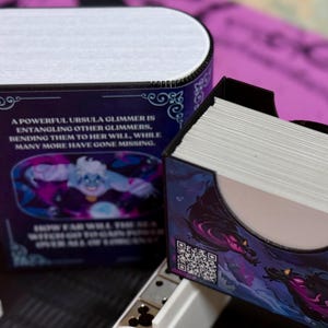 May include: A purple and blue deck box with a black and white illustration of a sea creature. The box has a quote on the front that reads "A powerful Ursula Glimmer is entangling other Glimmer, bending them to her will, while many more have gone missing."