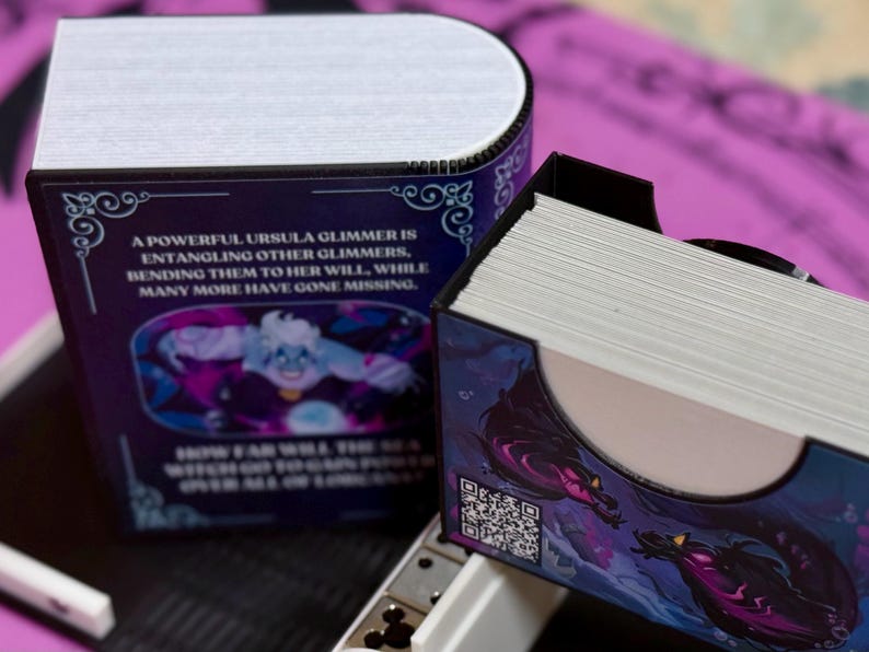 May include: A purple and blue deck box with a black and white illustration of a sea creature. The box has a quote on the front that reads "A powerful Ursula Glimmer is entangling other Glimmer, bending them to her will, while many more have gone missing."