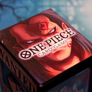 Op de afbeelding: Een close-up van een One Piece kaartspel doos. De doos toont een roodhuidig ​​personage met een kruisvormig litteken en het logo van het spel. De woorden "ONE PIECE CARD GAME" zijn in het wit gedrukt.