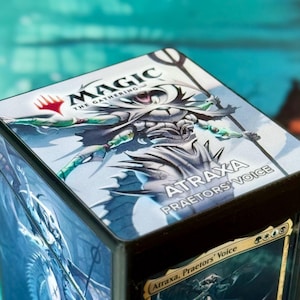May include: Black card storage box featuring artwork and text from Magic: The Gathering. The box displays the text "MAGIC THE GATHERING" and "ATRAXA PRAETORS' VOICE". The box has a black frame and a colorful illustration.