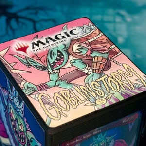 Peut inclure: Boîte de cartes Magic: The Gathering avec une illustration de gobelin et le texte "GOBLINSTORM" en jaune. La boîte a une palette de couleurs rose et bleu, avec le logo Magic: The Gathering en haut.