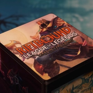 Può includere: Una scatola di carte da gioco con la scritta "RIFTBOUND LEAGUE OF LEGENDS TRADING CARD GAME". La scatola presenta un'illustrazione colorata di personaggi del gioco. La scatola è nera con una parte superiore colorata.