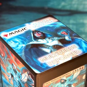 May include: A square deck box featuring Magic The Gathering branding with the text Reality Fracture and Multiverse Reforged. The top surface displays a hooded figure in white and blue robes surrounded by glowing blue energy, with Jace, Multiverse Architect visible on the side.