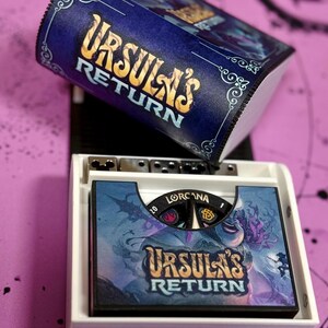 May include: A white box with a blue and gold lid. The lid has the text "Ursula's Return" on it. The box contains a game board with the same text and a character illustration. The game board has a black and white dial with the text "Lorcaana" on it.