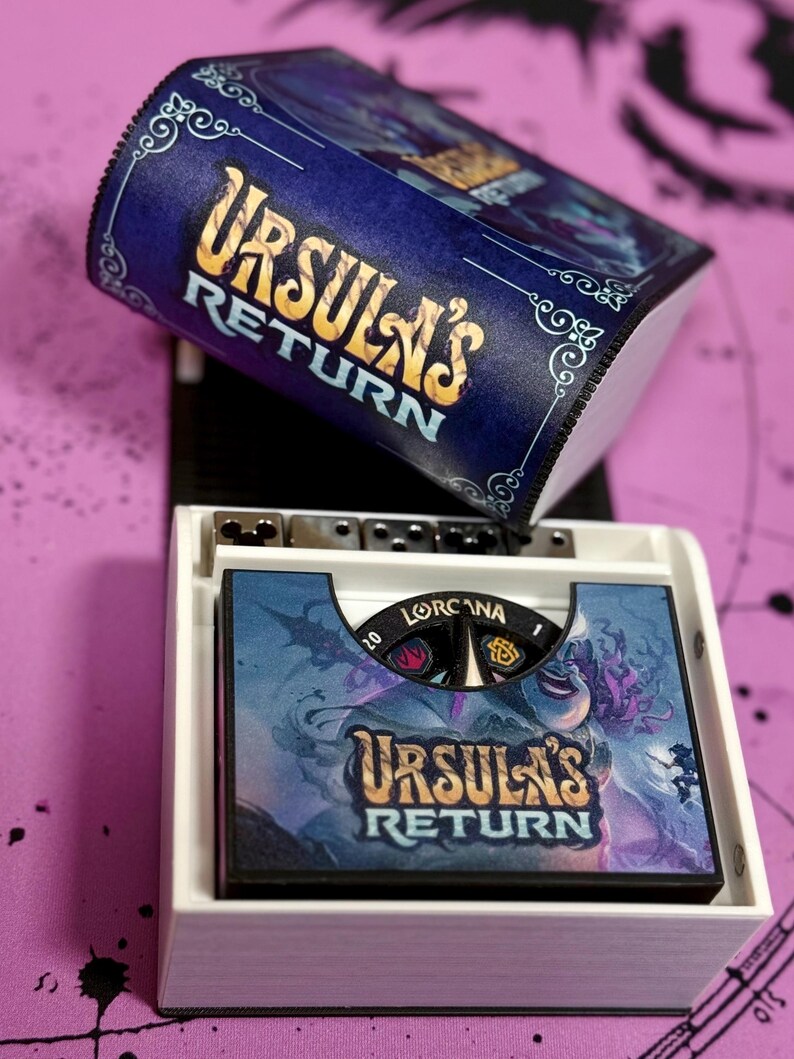 May include: A white box with a blue and gold lid. The lid has the text "Ursula's Return" on it. The box contains a game board with the same text and a character illustration. The game board has a black and white dial with the text "Lorcaana" on it.