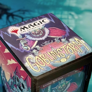 Peut inclure: Une boîte de jeu noire pour cartes à collectionner avec le logo Magic The Gathering et une illustration colorée d'un chef gobelin portant le texte GOBLINSTORM. Le côté de la boîte montre une vue partielle d'une carte Krenko, Mob Boss.