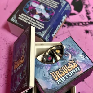 May include: A closed box with a blue and purple design featuring the text "Ursula's Return". The box is open to reveal a smaller box with a black and white spinning wheel with the text "Lorcaana" and "20".