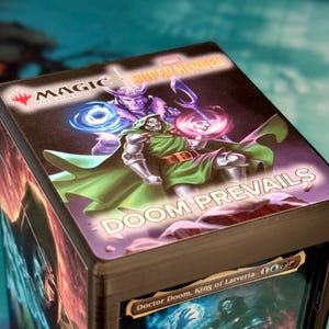 Op de afbeelding: Een rechthoekige deckbox voor ruilkaarten met een afbeelding van Doctor Doom en Loki uit Marvel Super Heroes. Het bovenoppervlak toont de tekst Magic The Gathering, Marvel Super Heroes en Doom Prevails. Een kaart is binnenin zichtbaar.