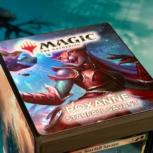 May include: A square Magic The Gathering deck box featuring a colorful illustration of a cat-like character in red armor against a cosmic space background. The text Magic The Gathering is at the top, and the name Roxanne Starfall Savant is below.