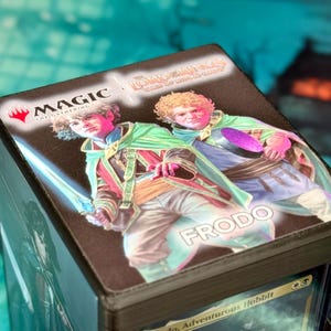May include: A black deck box for trading cards featuring a graphic of Frodo from The Lord of the Rings: Tales of Middle-earth. The top displays the Magic: The Gathering logo, the title text, and two illustrations of Frodo wearing green cloaks.
