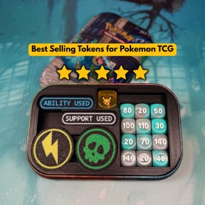 Könnte beinhalten: Ein schwarzes, rechteckiges Organizer-Tablett mit Pokemon TCG-Spielzubehör. Es enthält zwei rechteckige Marker mit der Aufschrift ABILITY USED und SUPPORT USED, zwei runde Spielsteine mit Blitz- und Totenkopf-Symbolen sowie zwölf Würfel.