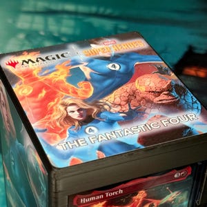 Può includere: Un contenitore per mazzi quadrato con il marchio Magic The Gathering e Marvel Super Heroes, che raffigura i personaggi dei Fantastici Quattro, tra cui la Torcia Umana, la Donna Invisibile, Mr. Fantastic e la Cosa.