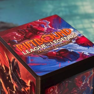 May include: A black box featuring the "Riftbound League of Legends Trading Card Game" logo in orange and white. The box has a red and blue design with illustrations of characters from the game.
