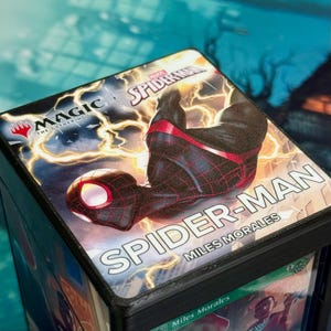 Puede incluir: Una caja negra para guardar cartas con un diseño de Spider-Man. La caja tiene las palabras "SPIDER-MAN" y "MILES MORALES" impresas. La parte superior de la caja tiene un gráfico de Spider-Man con un fondo de rayos. La caja también tiene el logotipo de "MAGIC THE GATHERING".
