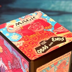 Puede incluir: Una caja cuadrada para guardar cartas Magic The Gathering Secret Lair Odds and Ends con una vibrante ilustración multicolor de una figura con cuernos y tres ojos en la tapa, sobre un fondo azul brillante con estrellas amarillas.