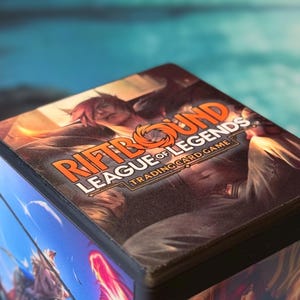 May include: A stack of "Riftbound League of Legends" trading cards. The box has a character illustration and orange and white text. The background is a blurred image of a blue and teal sky.