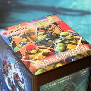 May include: A square deck box featuring Teenage Mutant Ninja Turtles artwork on the top lid with the text Magic The Gathering and Turtle Power. The side panel displays a Magic The Gathering card titled Leonardo, the Balance.