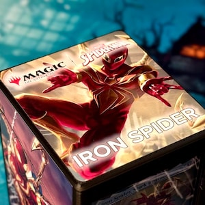 Puede incluir: Caja de cartas Magic: The Gathering con Iron Spider de Marvel. La caja es roja y dorada, con el personaje Iron Spider en una pose dinámica. El texto "IRON SPIDER" se muestra de forma destacada.